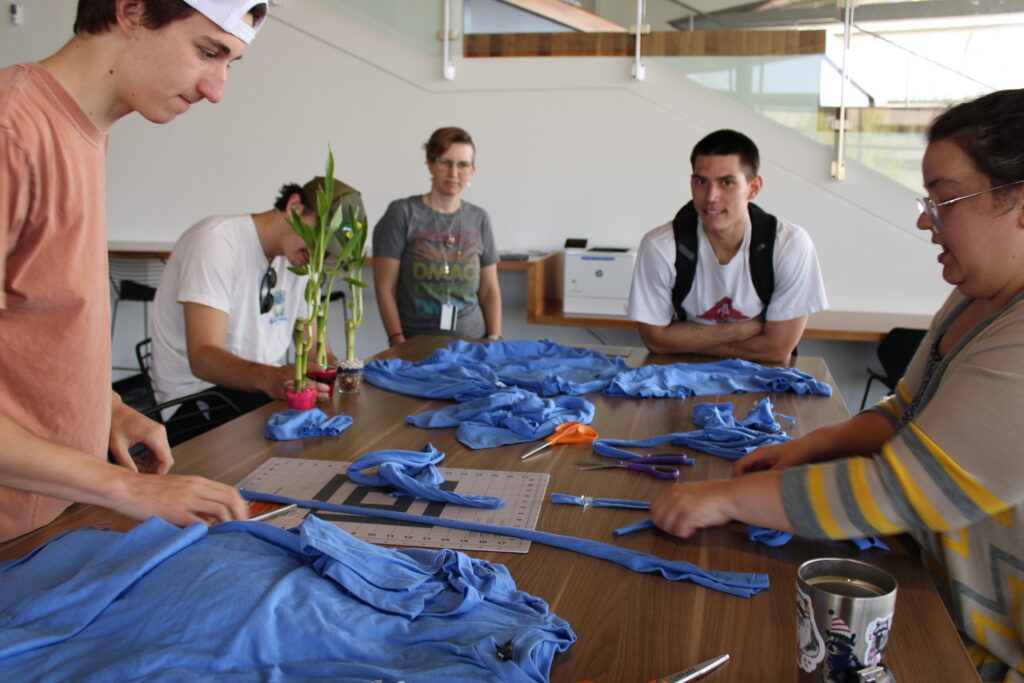 Students and staff surround a table full of crafting supplies and T-Shirt scraps at a Earth Day station. 