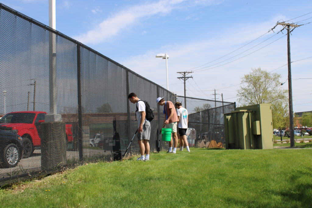 Students are stood up against a fence with buckets and grabby tools picking up trash at Des Moines Area Community College. 
