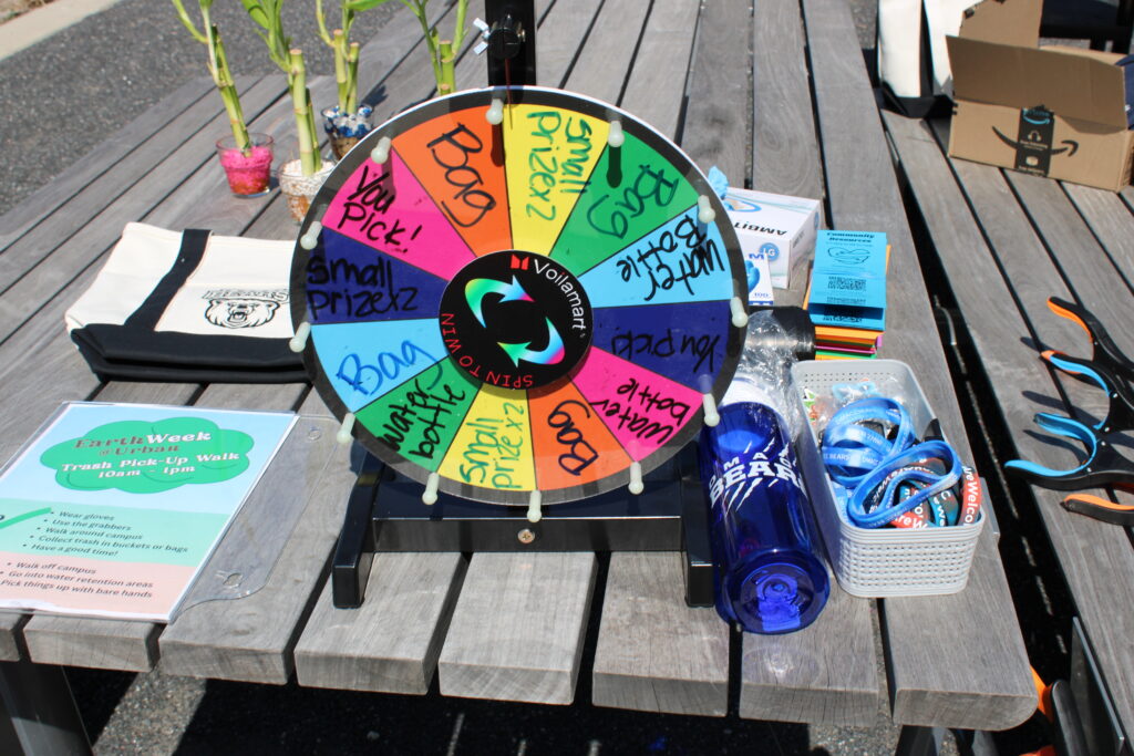 There is a picnic table with various Des Moines Area Community College merchandise surrounding a spinner wheel. Water bottles, bracelets, bookmarks, and book bags are showcased as prizes. 