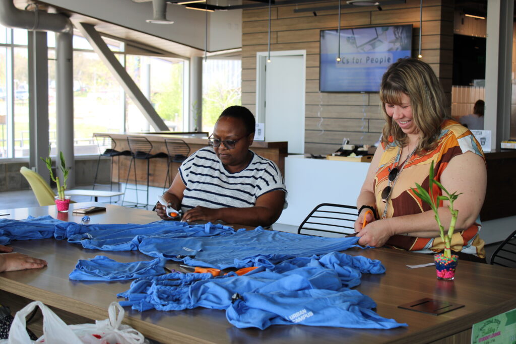 Two women sit side by side at a table cutting fabric. 