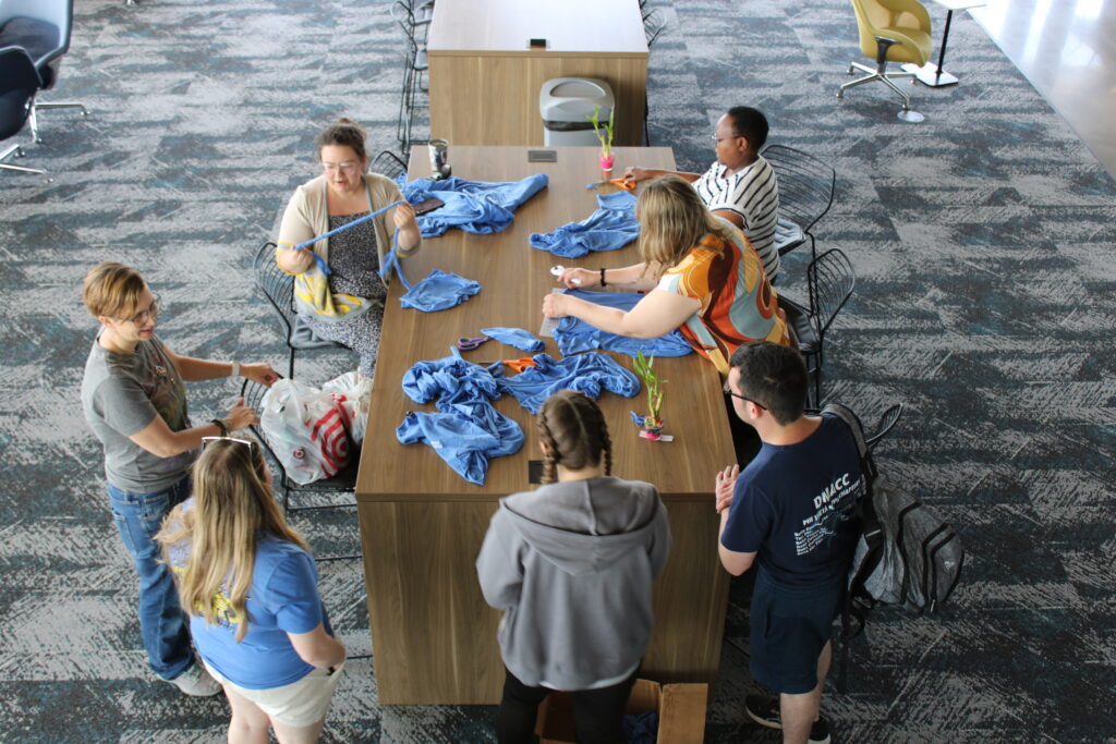 Three people sit at a table that has lots of crafting supplies on it. One gives a demonstration to four onlookers. 
