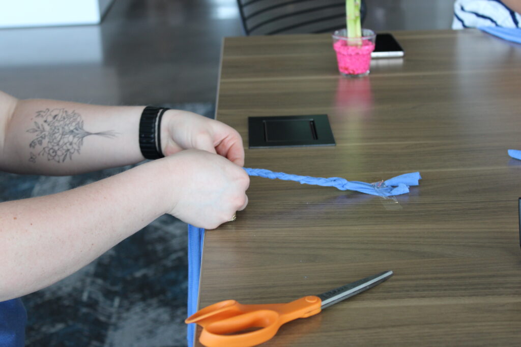 There are two hands coming from the left side of the photo grasping fabric. They are being braided together. There are scissors and a cup with a bamboo plant also pictured.